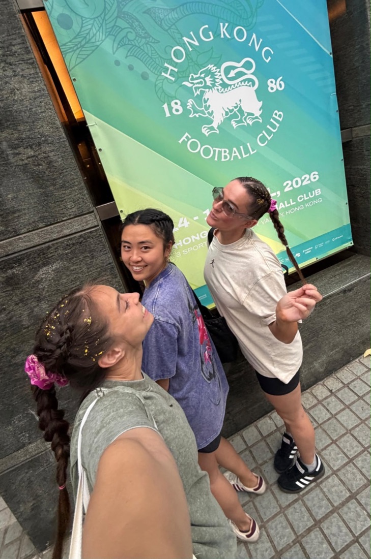 Three of the team in front of the Hong Kong Football Club banner, all wearing braids